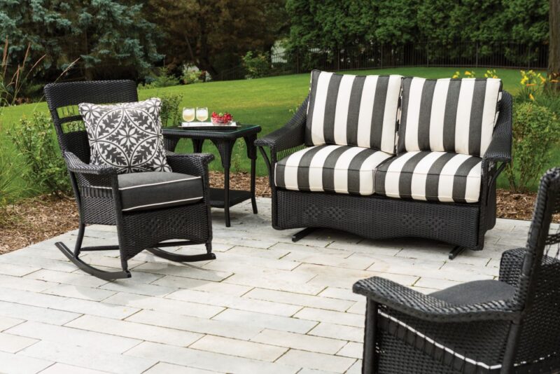 Outdoor patio set with a striped sofa, two chairs, and a small table, set on a stone patio surrounded by a lush green garden with an inserted fire pit.