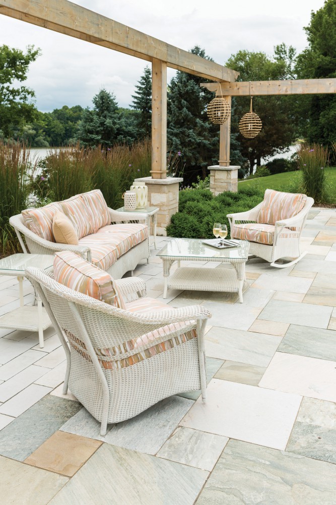 An inviting outdoor seating arrangement featuring white woven chairs with orange-striped cushions, a small table with two glasses of wine, set on a stone patio overlooking a lush landscape and warmed by a nearby fire pit.