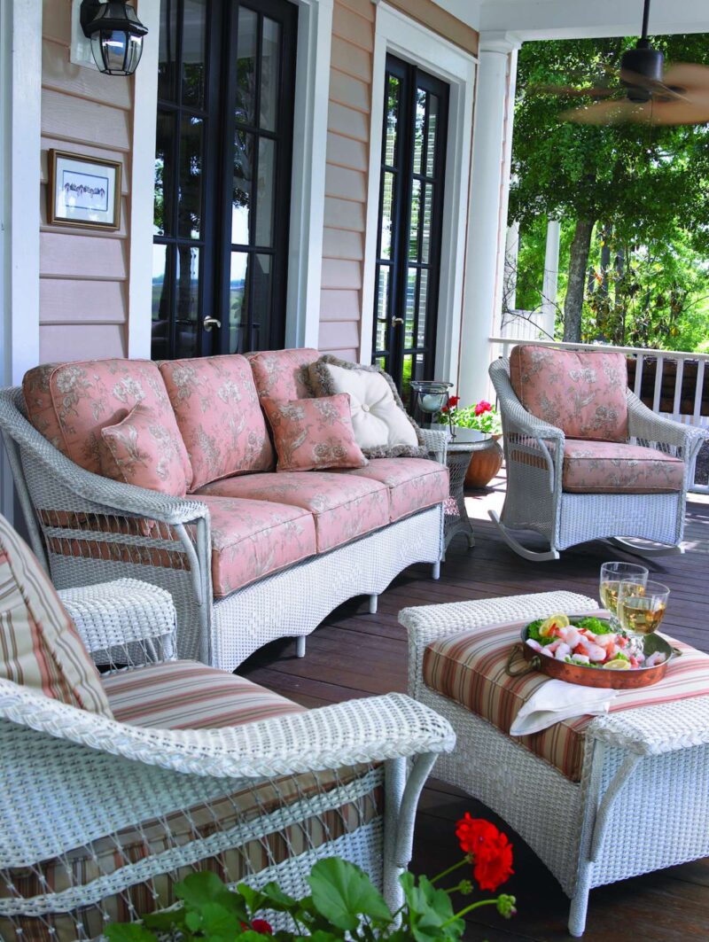 A cozy, well-furnished porch with wicker furniture featuring a sofa and chairs, a coffee table with a platter of fruits and a glass of wine. Vibrant flower pots and a stylish fire