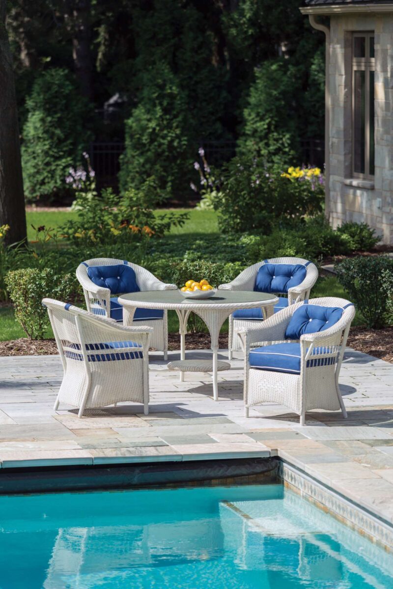 Elegant outdoor seating area by a pool, featuring four white wicker chairs with blue cushions around a coffee table with a fruit bowl and a fire pit, set in a lush garden.