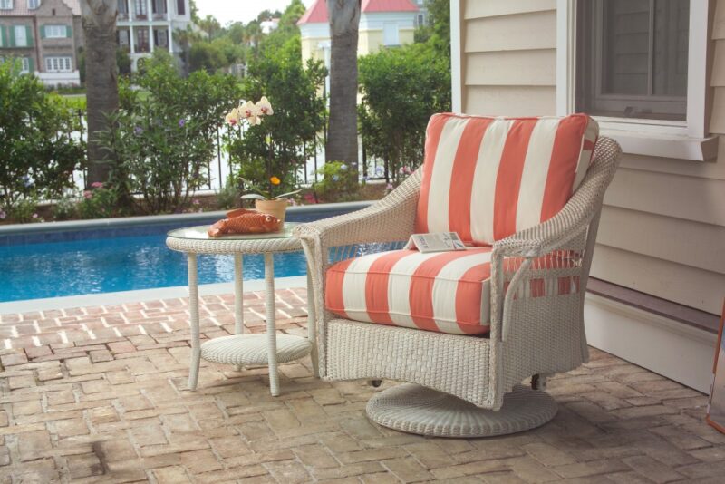 A cozy outdoor seating area with a cushioned wicker chair and matching table, set near a pool. On the table are a book, a cup, and a woven basket, with colorful houses in