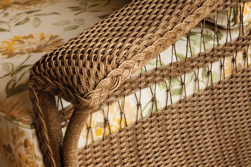 Close-up of a wicker chair armrest with intricate weaving details, positioned against a cushion with a floral pattern insert.