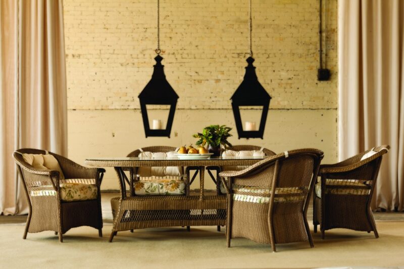Elegant outdoor dining setting with a rattan table and chairs, stylish lanterns hanging above, against a rustic brick wall backdrop, adorned with a centerpiece of fresh fruits and a chic fire pit nearby.