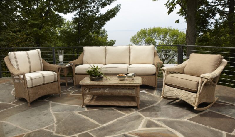 Outdoor patio furniture set on a stone terrace, featuring a beige sofa, two armchairs, and a coffee table with an integrated fire pit, overlooking a blurred green and blue backdrop.