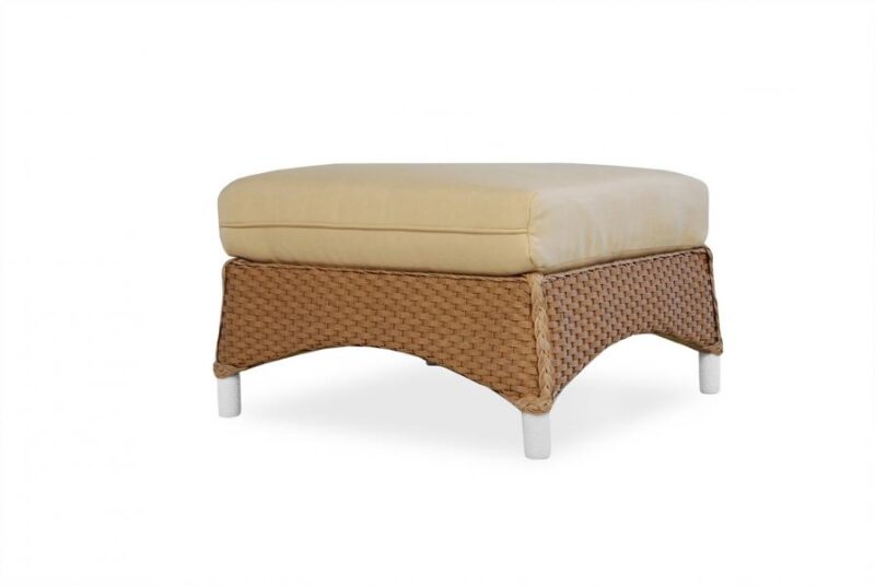 A wicker ottoman with a cream cushion near a fireplace, set against a white background. The ottoman has a rectangular base and short, sturdy legs.