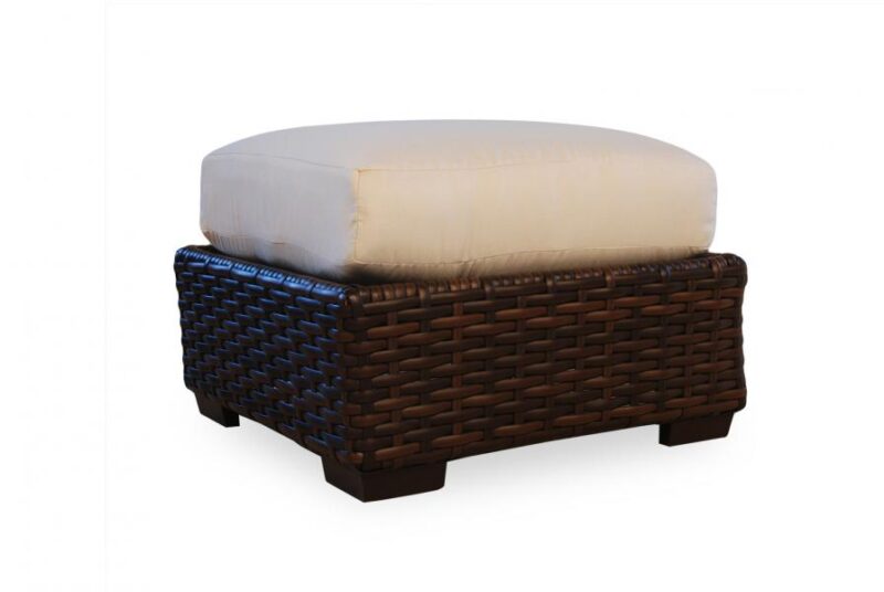 An ottoman with a cushioned beige top and a dark brown woven wicker base, set against a plain white background near a fireplace.
