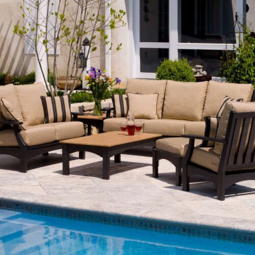 An elegant outdoor patio set with cushioned chairs and a coffee table beside a swimming pool, in a sunny backyard setting, includes an inviting fireplace.