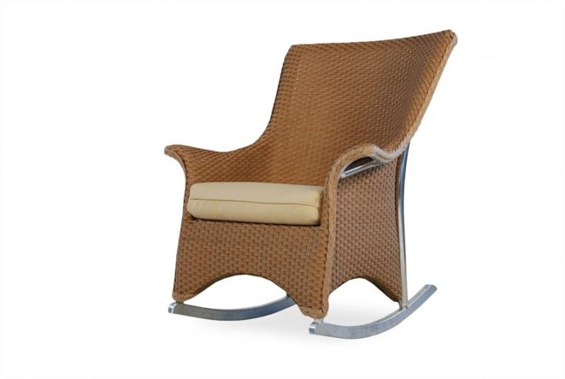 A brown wicker rocking chair with a beige cushion, set against a white background. The chair features curved armrests and a high back, resting on a smooth, light metal base near a fireplace