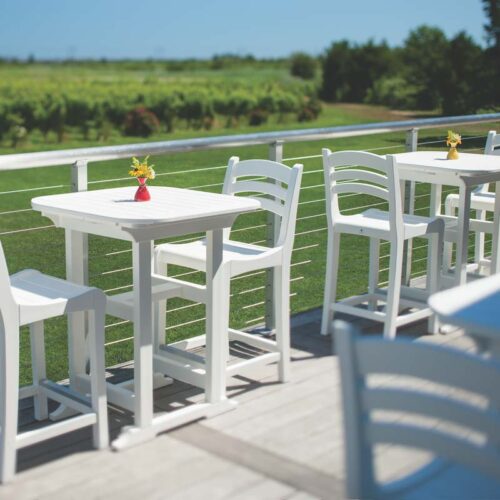 Outdoor patio with white tables and chairs overlooking a scenic green vineyard on a sunny day, each table adorned with a small yellow flower in a vase and an elegant fireplace nearby.