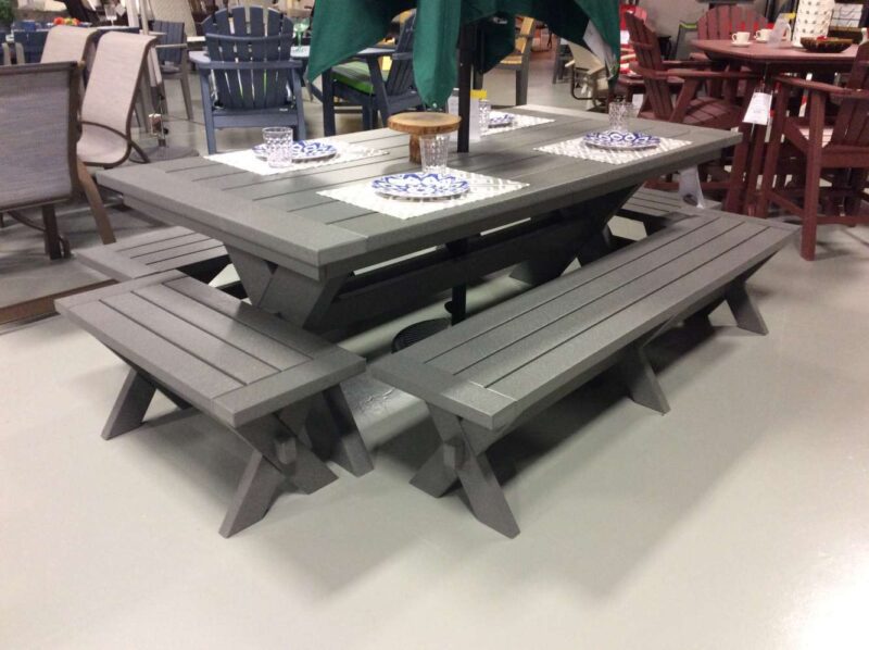 A gray outdoor picnic table with attached benches, displayed in a store. The table is set with two decorative mosaic coasters and features a built-in fireplace. Various other patio furniture is visible in the background