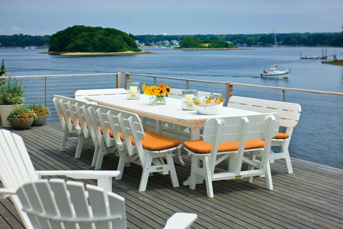 Outdoor dining setup on a wooden deck overlooking a scenic river view, featuring a long white table with orange-cushioned chairs, vases with yellow flowers, and a cozy fire pit nearby, with a