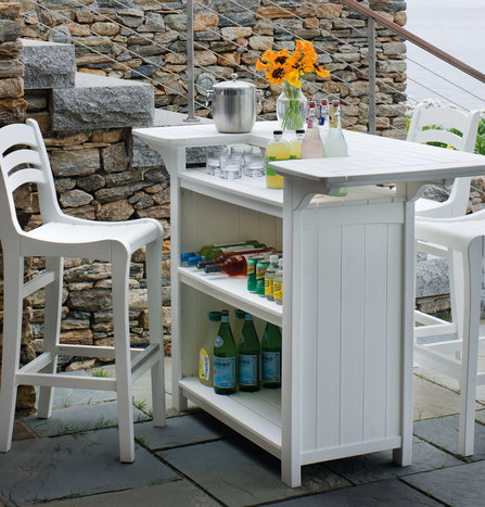 An outdoor bar setup with a white serving cart stocked with drinks and snacks, next to white chairs and a stone wall, overlooking a serene environment with an inserted fire pit.