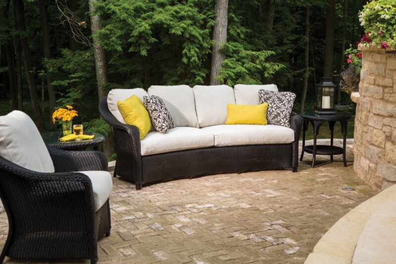 An elegant outdoor patio setting in a lush garden, featuring a corner sofa with white cushions, black wicker frames, and patterned yellow throw pillows. Nearby are a single armchair and a stone table