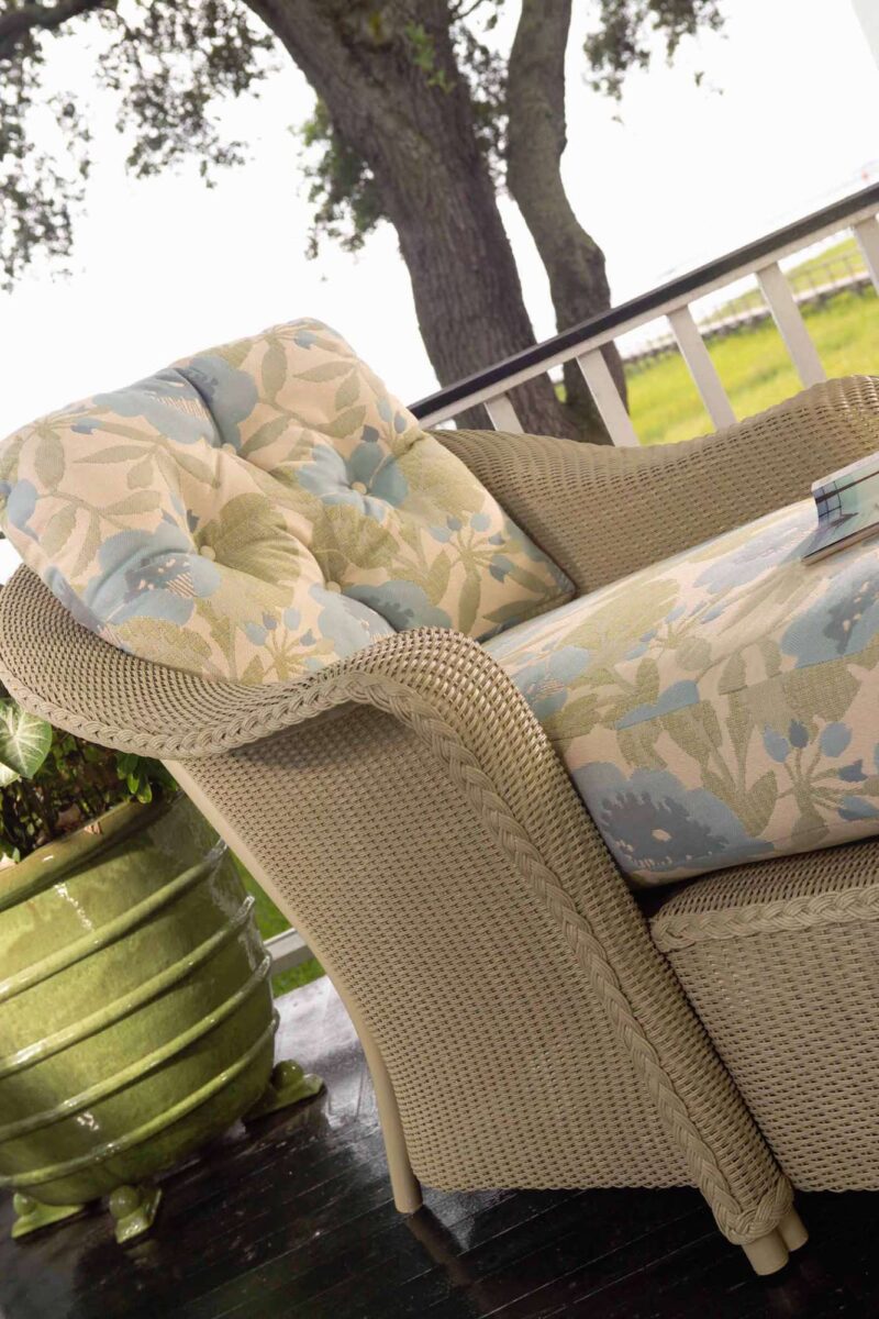 A cozy wicker armchair with floral cushions on a porch, overlooking a lush garden. A decorative fire pit is beside the chair.