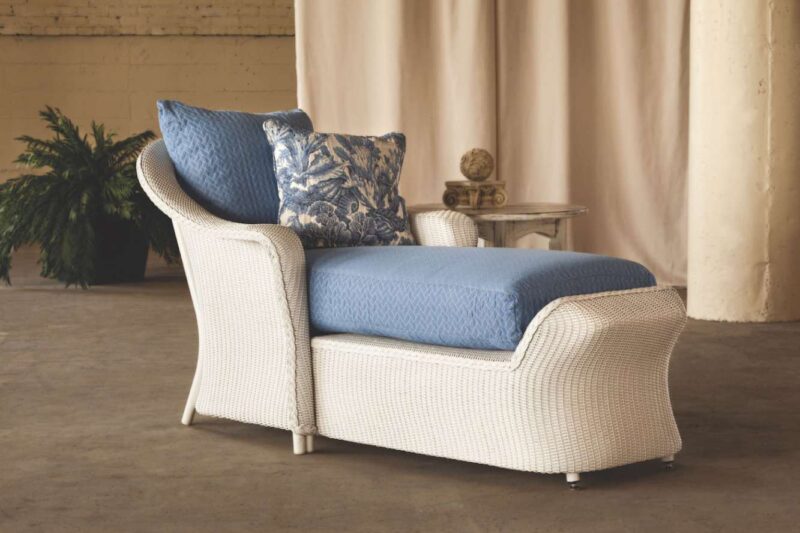 A stylish chaise lounge with a textured white frame and blue cushions, with a matching blue and white pillow. It's set against a backdrop of a creamy curtain and a side table with a plant,