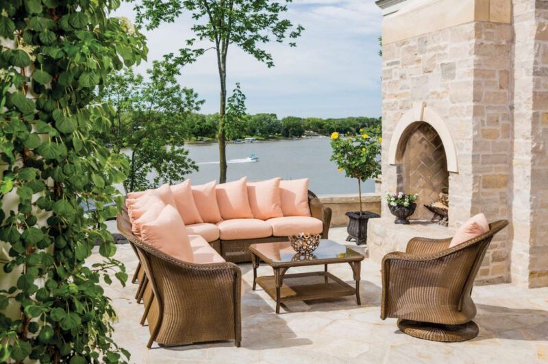 An elegant outdoor patio with wicker furniture and soft pink cushions, overlooking a serene lake. A stone fire pit adds charm to the tranquil setting.