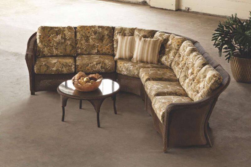 A wicker sectional sofa with floral cushions arranged in an L-shape around a small glass-topped coffee table, placed in a room with a concrete floor, a fireplace, and a potted plant