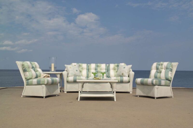 Outdoor patio furniture arranged on a beach, consisting of a sofa and two chairs with green cushions, and a coffee table with a book and drinks, under a clear blue sky, near an insert fireplace.