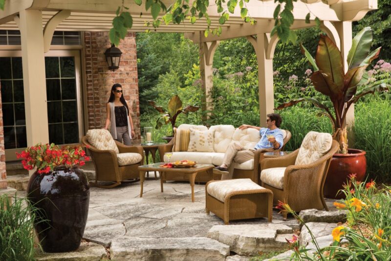 An outdoor patio setting with three people relaxed on wicker furniture, surrounded by lush greenery and flowers, with a brick house featuring an insert fireplace in the background.