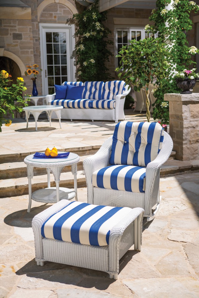White and blue striped outdoor furniture set on a stone patio, surrounded by a stone house and green plants, with a fire pit accentuating the colors on a bright day.