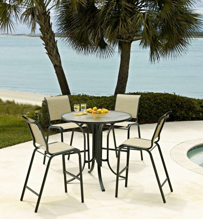An outdoor dining setup near a pool, featuring a metal table with four chairs and fruit on the table, with palm trees and a fire pit in the background.