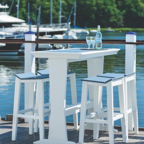 White high-top table with two stools set on a dock, overlooking a marina with boats. A bottle and two glasses sit on the table, next to an elegant fireplace insert, suggesting a relaxing scene