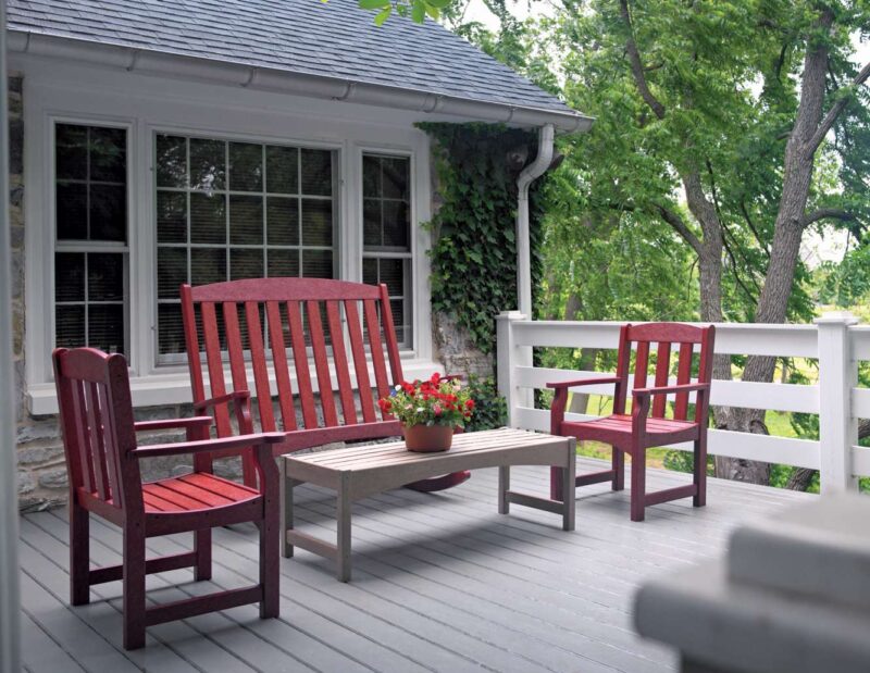 A cozy porch with two red adirondack chairs, a side table with red flowers, and a small stove, overlooking a lush garden, offering a quiet, relaxing outdoor space.