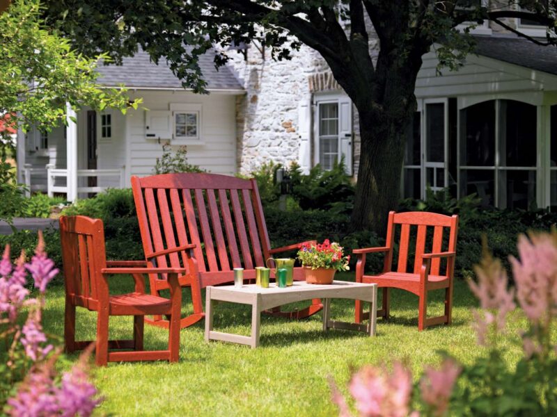 Two red wooden adirondack chairs and a small table with drinks and a potted plant, set on a lush lawn near a white house with visible stonework and an inviting fire pit.