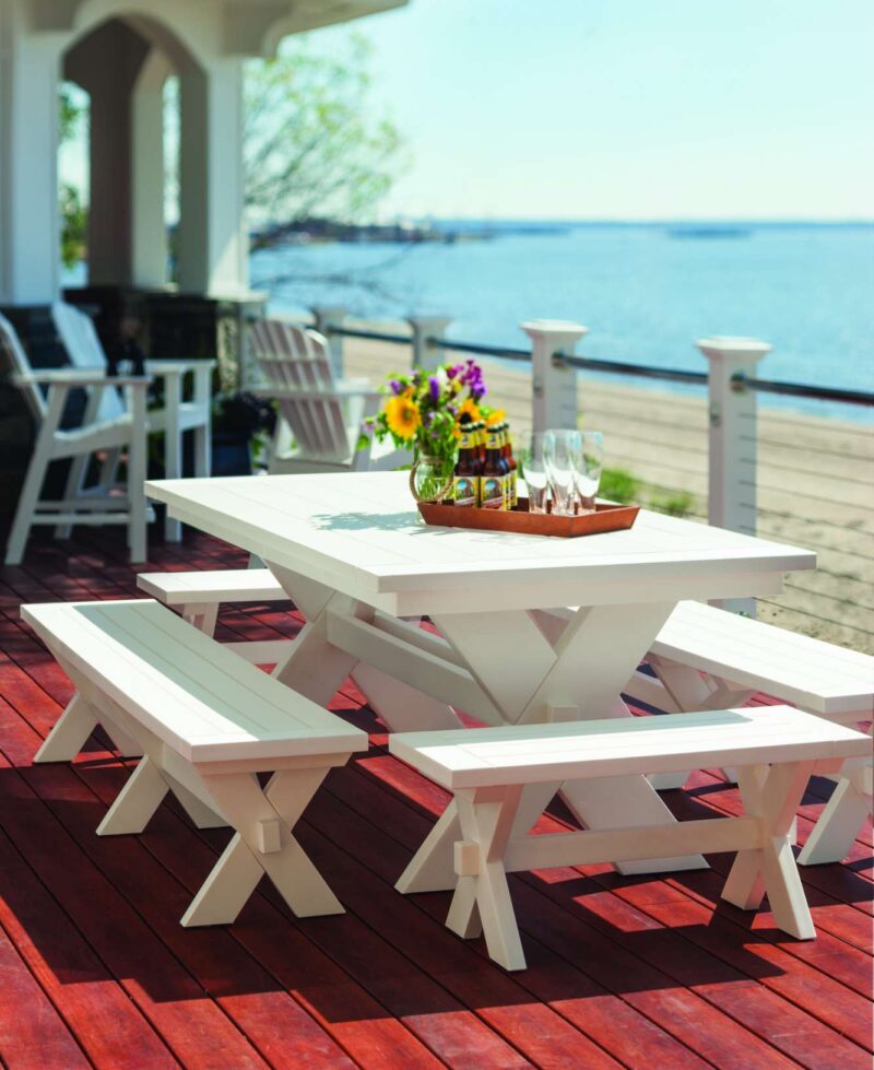 A scenic outdoor setting with a white picnic table set on a red wooden deck. The table is adorned with a centerpiece of colorful flowers, overlooking a tranquil blue sea under a clear sky. Nearby, an
