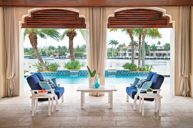 A luxury outdoor seating area featuring two blue chairs with white frames facing a pool, framed by large arch windows with sheer curtains and a view of palm trees, a waterway, and an elegant fire pit
