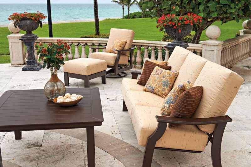 An elegant outdoor seating area with a plush beige sofa and chairs, a brown coffee table, decorative cushions, and a scenic view of a lush green lawn and ocean in the background, complete with a cozy