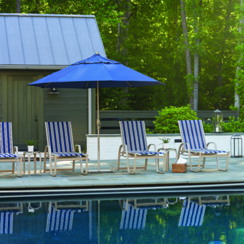 A serene outdoor pool area with four blue-striped lounge chairs under a blue umbrella, beside a pool reflecting the surrounding green trees. A modern gray pool house with an elegant fireplace complements the tranquil setting.