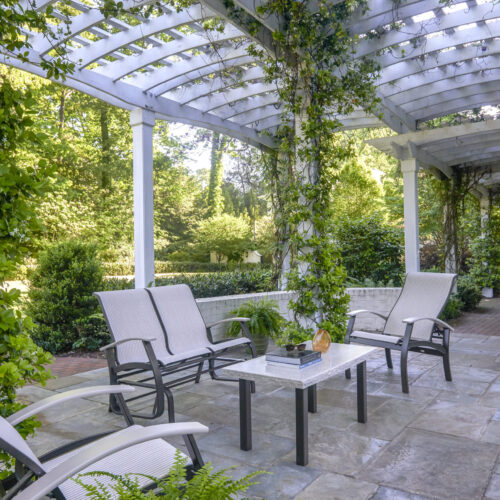 A serene outdoor seating area under a white pergola covered with climbing green ivy, featuring four sleek lounge chairs around a small coffee table with an integrated fire pit, surrounded by lush greenery.