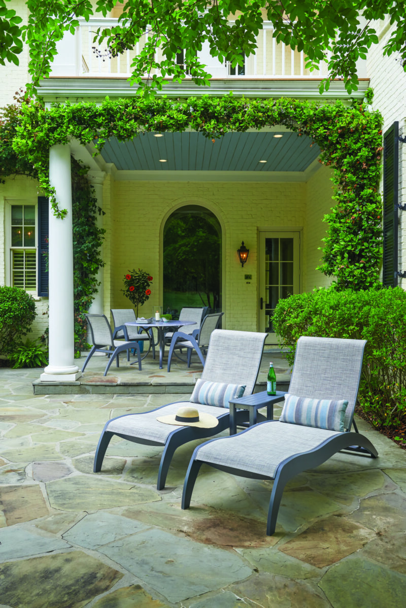 Two lounge chairs and a small table with a book and drink are positioned on a stone patio under a covered archway, next to an outdoor stove, surrounded by lush ivy and a traditional white brick