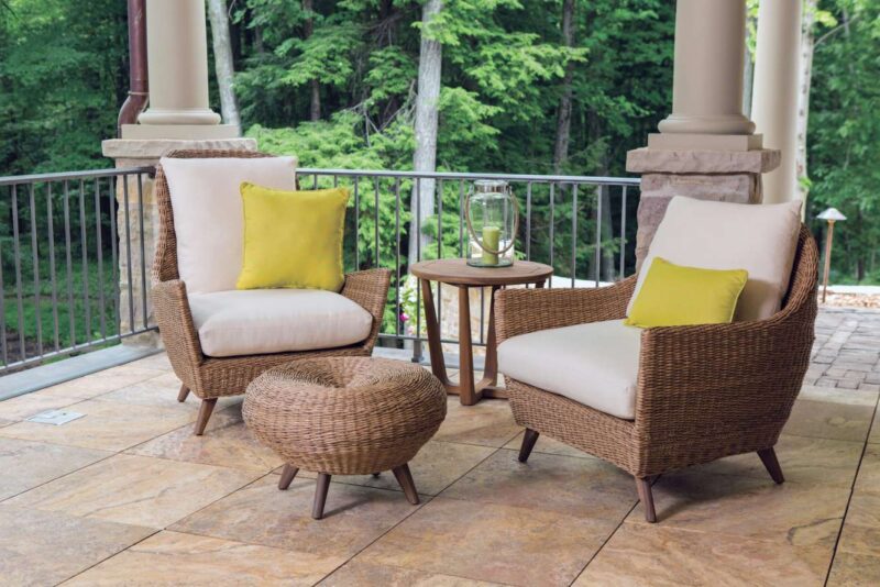 Two wicker chairs with white cushions and yellow pillows on a tiled patio, a small round wicker table between them, overlooking lush green woods near an outdoor fireplace.