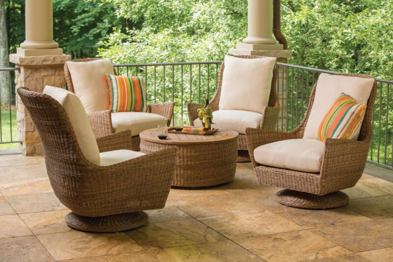 Four cozy, wicker chairs with white cushions and colorful stripes, arranged around a small table with a fire pit, on an elegant, covered patio overlooking greenery.