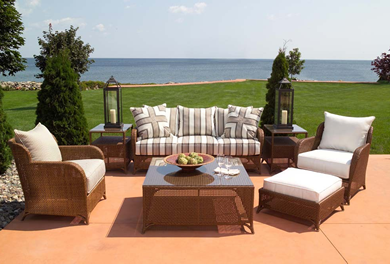 Outdoor patio furniture set on a terrace overlooking a lake under a clear blue sky, with matching sofa, armchairs, and a coffee table accented by a large fireplace and lanterns.