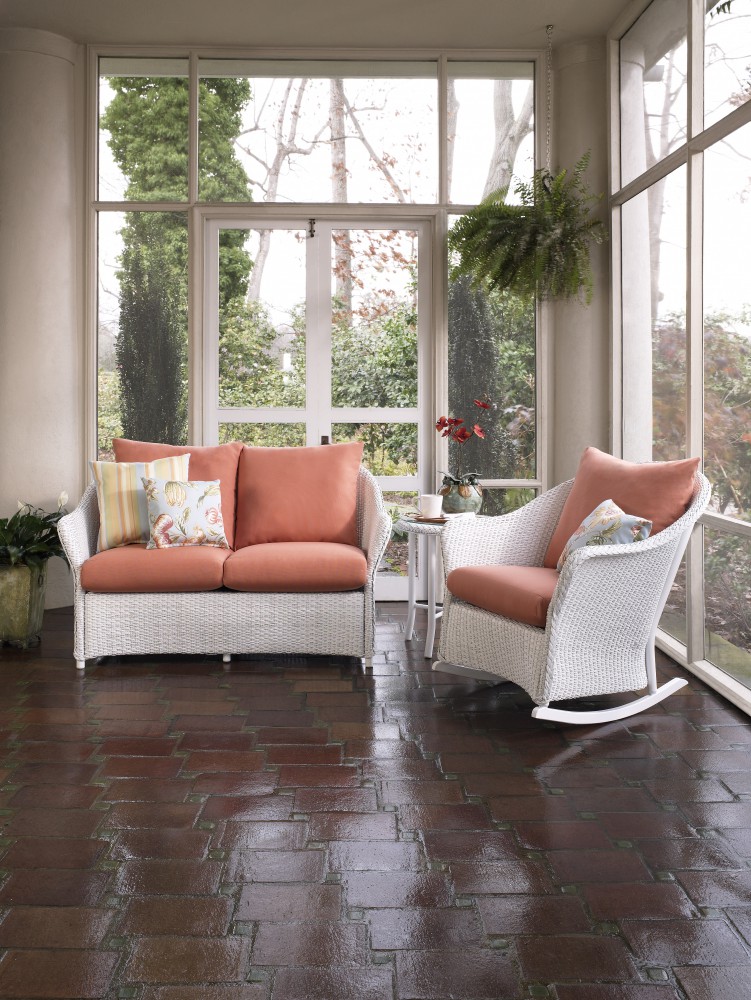 A cozy sunroom with large windows showing a garden view, furnished with a white wicker sofa and rocking chair with orange cushions, and a side table beside a fireplace, on a terracotta tiled