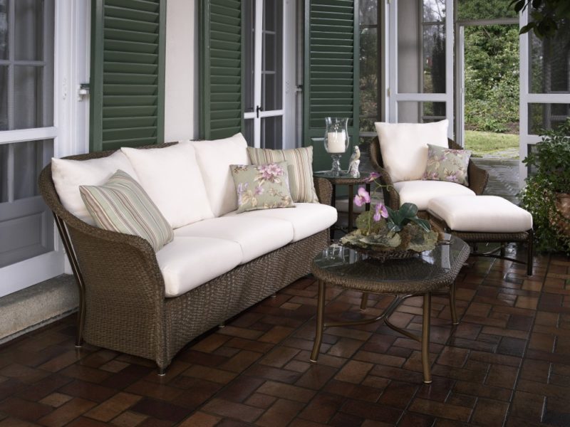 Outdoor patio furniture set with a wicker sofa, two matching armchairs, and a glass-top coffee table beside a fire pit on a brick floor, next to a home with green shutters.