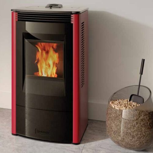 A modern pellet stove in red and black, with visible flames inside, is placed in a room corner near a glass container filled with wooden pellets.