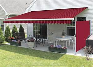 A spacious backyard featuring a red-striped awning extended over a patio with a dining set and a pizza oven, surrounded by neatly landscaped grass and shrubs near a white house.