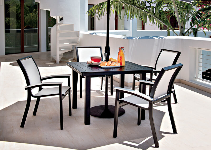 A modern outdoor dining setup on a white patio featuring a black table with four chairs, and breakfast items including a bottle of juice, a bowl of fruits, and an insert grill.