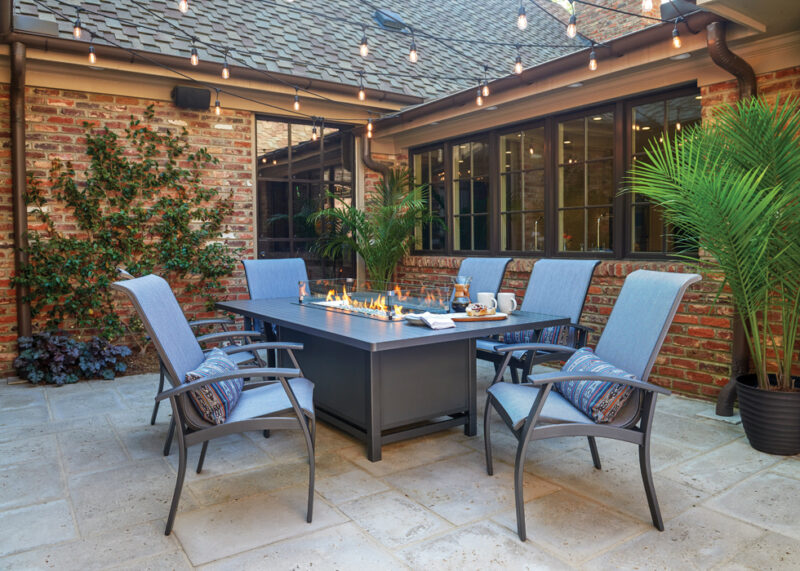 An inviting outdoor patio area with a dining table featuring a built-in grill, surrounded by chairs and decorative plants, with brick walls and strung lights above.