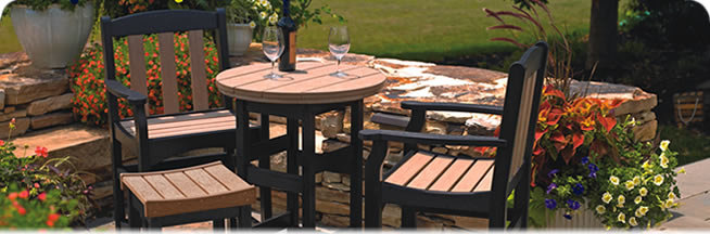 A cozy patio set up with a round counter collection table and four chairs made of dark wood, set on a stone paved area surrounded by lush plants and a short stone wall. Two wine glasses sit on