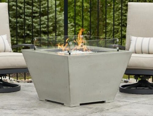 Two beige outdoor swivel chairs with cushions flank a modern square fire pit insert with dancing flames, set on a patio with a green hedge background.