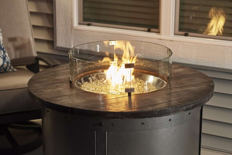A modern gas fireplace table with a circular glass wind guard and golden fire glass, set on a wood-finished outdoor patio near a window reflecting flames.