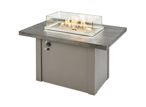 A modern outdoor fireplace table with a glass wind guard and a visible flame, set on a gray, stone-textured top and a taupe-metal base.