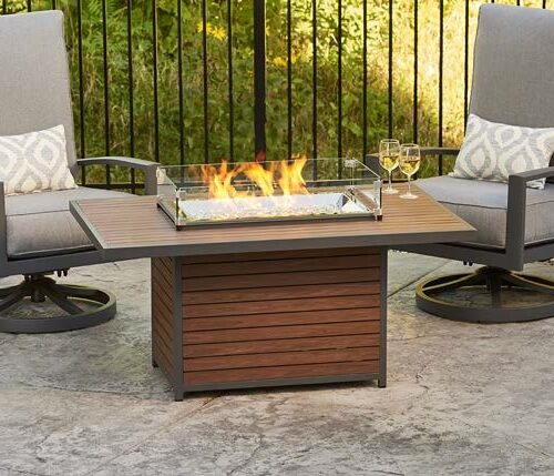 A cozy outdoor seating area featuring two chairs with grey cushions flanking a fire pit insert with a glass shield, a glass of wine on the table, set on a patterned patio surface.
