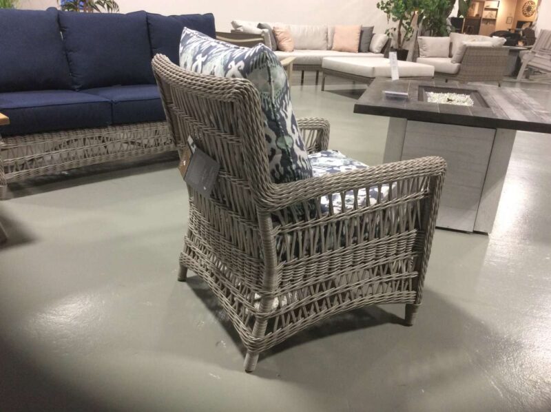 A grey wicker chair with blue and white patterned cushions displayed in a furniture store with other sofas, chairs, and a fire pit in the background.