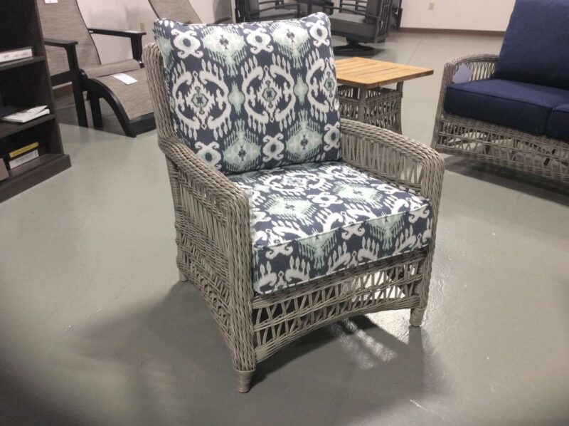 A wicker armchair with a white and blue patterned cushion set in a furniture showroom with other chairs, tables, and a fireplace in the background.